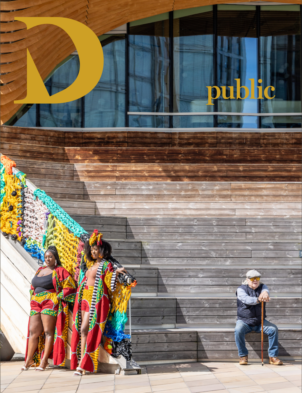 A building in London's Olympic Park, two young women pose for an unseen photograph in colourful outfits. To the right, an older person with cane sits on the wooden steps. 